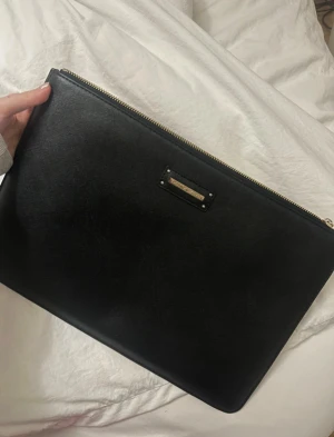 Svart kuvertväska från ideal of Sweden  - Stilren svart kuvertväska från ideal of sweden med guldfärgad dragkedja och metallplakett framtill. 