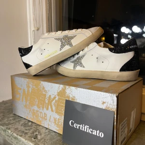 Golden goose Superstar med glitter silverstjärna - Säljer ett par vita Golden Goose sneakers med silverglittrande stjärna på sidan och svart glittrig häl. Skorna har snörning, platt sula och GGDB/SSTAR-tryck i guld. Perfekta för dig som gillar coola detaljer och unik design.