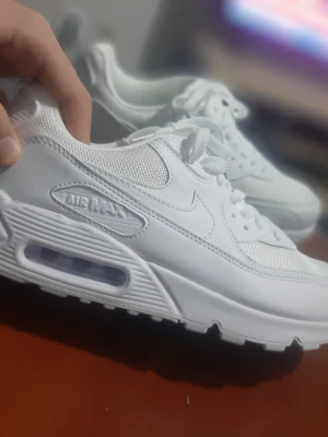 Nike Air Max 90 helvita sneakers - Nike Air Max 90 sneakers i helvit färg med mesh och syntet på ovandelen, klassisk Air Max-dämpning i sulan och tydlig logga på sidan. Skorna har snörning och en rund tå, perfekt för dig som gillar stilrena och sportiga sneakers.