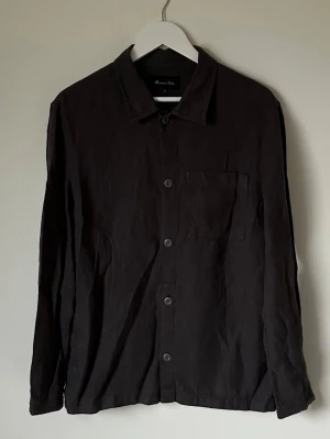 Mörkgrå linné overshirt från Massimo Dutti - Mörkgrå overshirt från Massimo Dutti i 100% linne. Klassisk krage, knappstängning framtill och en bröstficka. Långärmad och med en clean, minimalistisk look som funkar till många stilar. Perfekt för dig som gillar enkla och stilrena plagg. 5L8