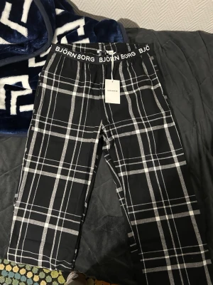 Björn Borg mjukisar  - Svarta byxor från Björn Borg med vitt rutmönster och bred resår i midjan med logotyp. Sköna och stilrena, perfekta för chill eller häng. Materialet är mjukt och känns som bomull. Klassisk passform och snyggt mönster som sticker ut.