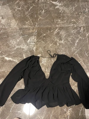 Svart omlottblus med volang - Säljer en svart blus med omlott-design och djup v-ringning. Blusen har långa ärmar och knytband i nacken. Volangdetaljer vid midjan ger en snygg och feminin siluett. Perfekt att styla med jeans eller kjol för en trendig look. Även väldigt fin till fest och fina middagar 