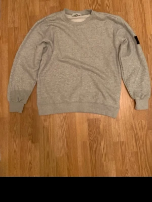 Grå sweatshirt från Stone Island - Grå sweatshirt från Stone Island med klassisk rund hals och ribbade muddar. Tröjan har lång ärm och den ikoniska Stone Island-loggan på vänster ärm. Materialet är mjuk bomull som ger en skön och avslappnad känsla. Perfekt för chill dagar och streetwear-stil.