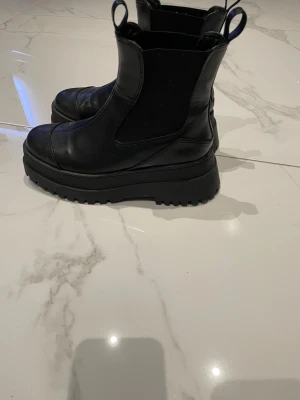 Svarta chunky chelsea boots - Säljer ett par svarta chelsea boots med chunky platt sula och grovt mönstrad yttersula. Skorna är tillverkade i skinn och har elastiska paneler på sidorna samt dragflikar fram och bak. Perfekta för dig som gillar en edgy stil och vill ha boots som sticker ut.