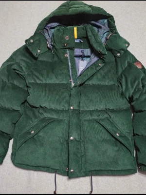 Grön manchester dunjacka Polo Ralph Lauren - Säljer en grön dunjacka från Polo Ralph Lauren i mjuk manchester. Jackan har huva, dragkedja och knappar framtill, samt stora fickor med tryckknappar. Insidan är fodrad med blågrönt rutigt mönster. Perfekt för kalla dagar! MÖTS BARA TYVÄR‼️‼️‼️