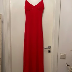 Röd långklänning med smala axelband - Snygg röd långklänning med smala justerbara axelband och dekorativ kant upptill. Klänningen har en figurnära passform och är tillverkad i ett mjukt, slätt material som ger ett elegant intryck. Perfekt för dig som vill sticka ut med en klassisk och stilren look. Storlek 34 men passar 36