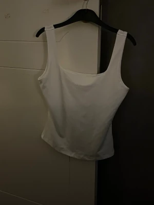 Vit linnetopp med breda axelband - En stilren vit linnetopp med breda axelband och rak halsringning. Toppen är figurnära och passar perfekt till jeans eller kjol. Gjord i mjukt material som känns skönt mot huden.