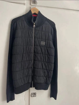 Svart quiltad bomberjacka från Hugo - Svart cardigan från Hugo med quiltad framsida och ribbstickade ärmar och krage. Jackan har en tydlig dragkedja framtill och en snygg logotyp på bröstet. Materialmix av polyester och stickad textil ger en cool och modern look.