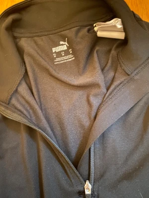 Svart långärmad träningströja Puma - Svart långärmad träningströja från Puma med halv dragkedja framtill och diskret mönster längs ärmarna. Tröjan har en stilren look med Puma-logga på bröstet och är gjord i ett mjukt syntetmaterial som andas. Perfekt för sport eller gym.