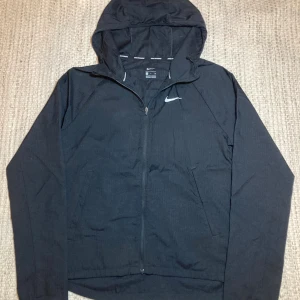 Svart Nike Miller vindjacka med huva - Nike vindjacka i svart med huva och dragkedja framtill. Jackan har ett diskret rutmönster i tyget och en vit Nike-logga på bröstet. Lätt och smidig design, perfekt för löpning och utomhusaktiviteter. Två sidofickor och sportig look. Reflexer på båda axlarna!