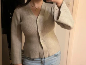 Beige ribbad kofta med knappar - Snygg beige kofta med ribbad struktur och v-ringad hals. Koftan har långa ärmar och stängs med fyra knappar framtill. Passformen är slim och materialet känns mjukt och stickat, perfekt för lager på lager under höst och vinter. Aldrig använd. 