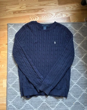 Mörkblå Ralph lauren stickadtröja - Kabelstickad mörkblå tröja från Ralph Lauren med klassisk rund hals och diskret broderad logga på bröstet. Tröjan har långa ärmar och ribbade muddar vid ärmslut och nederkant. Perfekt för dig som gillar stilren och tidlös design larger fit.