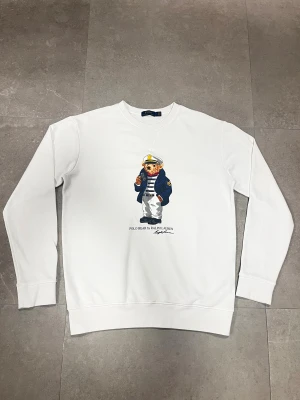 Vit sweatshirt Polo Bear Ralph Lauren - Vit sweatshirt från Ralph Lauren med ikoniskt Polo Bear-tryck på bröstet. Tröjan har rund hals, långa ärmar och ribbade muddar. Materialet är mjuk bomull som känns skönt mot huden. Perfekt för dig som gillar klassisk och lekfull stil.