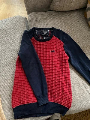 Röd och marinblå stickad tröja Bondelid - Säljer en snygg stickad tröja från Bondelid i storlek Large. Tröjan har röd kropp med strukturerat mönster och marinblå ärmar samt ribbade muddar. Liten broderad logga på bröstet. Perfekt för dig som gillar klassisk stil med en twist.