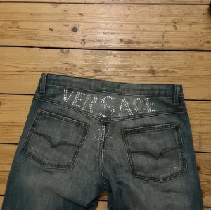 Versace jeans strass rhinestone  - Säljer ett par RARE blå jeans från Versace med låg midja. Baktill finns Versace-loggan i strass/rhinestone som glittrar ⭐️ Inköpta på Plick och bilderna är lånade från förra ägaren, men det är bara att skriva för fler bilder!! Dom passar mig som är ca 165 cm lång. Midjemåttet är ca 85, men det går att dra åt i och med snörning på insidan (bild 3) 