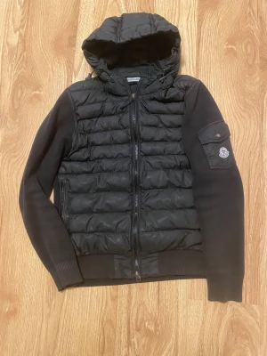 Svart pufferjacka med huva från Moncler - Svart pufferjacka från Moncler med quiltad front och huva. Jackan har stickade ärmar och rygg, dragkedja framtill och en ficka med Moncler-logga på ärmen. Perfekt för dig som vill ha en stilren och sportig look.