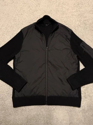 J.Lindeberg cardigan - En stilren och eftertraktad cardigan från J.Lindeberg, perfekt som ett lättare plagg till våren. Designad i klassisk svart färg med en diskret logga på armen som ger ett modernt och exklusivt uttryck. Enkel att matcha till både vardag och mer uppklädda outfits.  ✅ Märke: J.Lindeberg ✅ Storlek: L ✅ Färg: Svart ✅ Skick: Mycket bra ✅ Detaljer: Logga på armen  En snygg och tidlös J.Lindeberg-cardigan i mycket fint skick – redo att användas direkt.