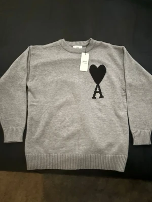 Grå stickad tröja från Ami Paris - Snygg grå stickad tröja från Ami Paris med svart hjärta och bokstaven A på bröstet. Tröjan har rund halsringning, ribbade muddar och är tillverkad i mjukt material. Perfekt för en stilren och trendig look. Priset kan diskuteras 