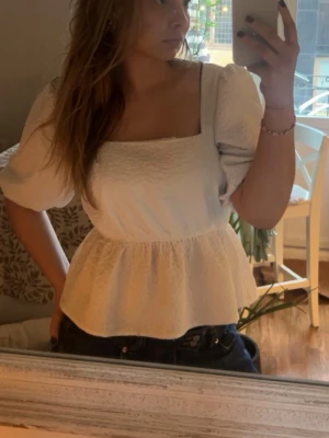 Vit puffärmad blus med fyrkantig ringning - Snygg vit blus med puffiga korta ärmar och fyrkantig ringning. Blusen har en lätt strukturerad yta och markerad midja med volang nedtill. Perfekt att styla med jeans eller kjol för en trendig look.