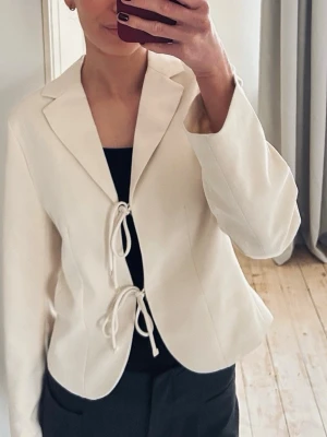 Vit kavaj med knytning fram - Stilren vit kavaj med två knytband framtill och klassisk krage. Modellen är något kortare och har en figurnära passform. Perfekt för en trendig och clean look. Storleken är Xs men passar även S. 🩷använd ca 4 gånger, köptes i december 2025! 