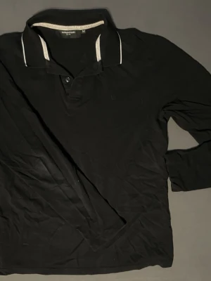 Svart långärmad piké från Lawrence Grey - Svart långärmad pikétröja från Lawrence Grey med klassisk krage och vita detaljer på kragen. Tröjan har knappar vid halsen och är tillverkad i ett mjukt bomullsmaterial. Perfekt för en stilren och avslappnad look.