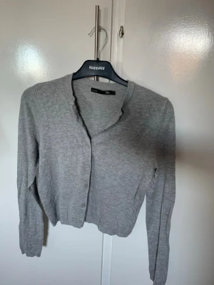 Grå kofta - Stilren grå kofta med v-ringning och knäppning framtill. Tillverkad i mjukt, tunt stickat material och har långa ärmar samt ribbade muddar vid ärmslut och nederkant. Perfekt att slänga över en topp för en enkel look.