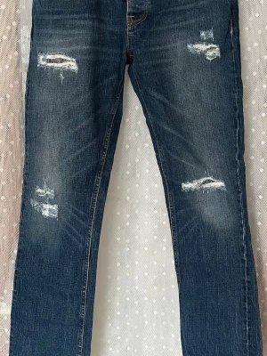 Blå slitna jeans från Nudie Jeans - Säljer ett par blå jeans från Nudie Jeans med snygga slitningar och hål på framsidan. Jeansen har klassisk femficksmodell, raka ben och kontrastsömmar på bakfickorna. Tillverkade i denim med en cool tvättad look. Grim Tim.  W33 L32
