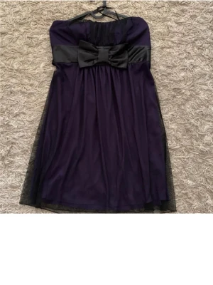 Mörklila axelbandslös klänning med rosett - Snygg mörklila axelbandslös klänning med svart satinband i midjan och stor rosett framtill. Klänningen har ett lätt utställt fall och är knälång med ett tunt lager tyll över kjolen. Perfekt för dig som vill sticka ut med en elegant look.