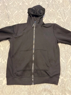 Svart hoodie från C.P. Company - Svart hoodie med dragkedja från C.P. Company. Utrustad med huva som har inbyggda glasögon, två stora fickor framtill och broderad logga på bröstet. Tillverkad i mjukt bomullsmaterial och har ribbade muddar vid ärmslut och nederkant.