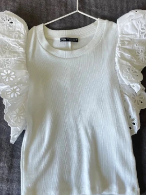 Zara fest topp med volanger  - Supersöt vit croppad topp från Zara med ribbad struktur och stora volangärmar i broderad spets. Perfekt för dig som vill ha en statement topp med unika detaljer och puffiga ärmar som verkligen sticker ut.