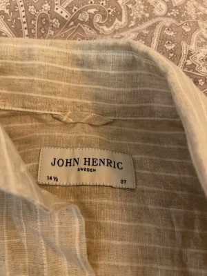 Beige randig linneskjorta John Henric - Snygg beige skjorta från John Henric i linne med tunna vita ränder. Skjortan har klassisk krage, knappar framtill och långa ärmar.