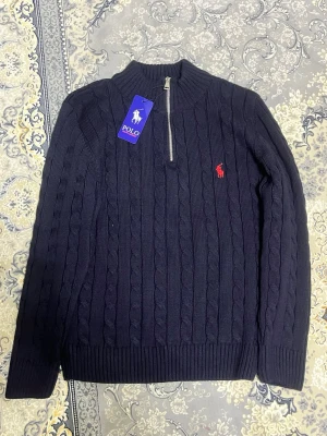 Mörkblå kabelstickad tröja Polo Ralph Lauren - Mörkblå kabelstickad tröja från Polo Ralph Lauren med halv dragkedja och röd broderad logga på bröstet. Tröjan har ribbade muddar och hög krage, perfekt för en stilren och klassisk look. Materialet känns mjukt och håller värmen.
