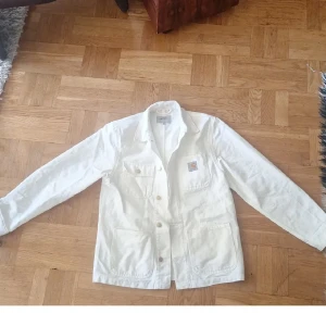 Vit overshirt från Carhartt - Vit overshirt från Carhartt med klassisk krage, fyra stora fickor framtill och knappar längs hela framsidan. Tillverkad i kraftig bomull och har en avslappnad passform. Perfekt för lager på lager och enkel att styla till olika outfits. Använd 2 gånger