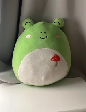 Grön groda Squishmallow med svamp - Supermjuk och rund grön Squishmallow i form av en groda med vita kinder och ett broderat rött svampmotiv på magen. Perfekt som  gosedjur. Den har söta öron och ett glatt ansikte.