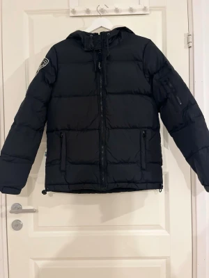 Svart dunjacka från D.Brand 90% dun - Svart dunjacka från D.Brand med 90% dun, broderad logga på ärmen, dragkedja framtill, två fickor med dragkedja och rymlig huva med dragsko. Perfekt för kalla vinterdagar och har en stilren, puffig design.