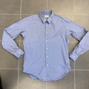 Blå randig skjorta från J.Lindberg  - Klassisk blå skjorta med tunna vita ränder från J.Lindberg . Skjortan har lång ärm, vit knappstängning och en stilren krage. Perfekt för en dressad look och tillverkad i ett lätt bomullstyg.