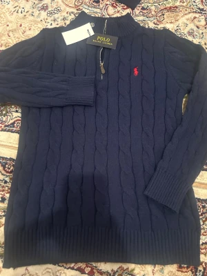Mörkblå kabelstickad tröja Polo Ralph Lauren - Mörkblå kabelstickad tröja från Polo Ralph Lauren med röd broderad logga på bröstet. Tröjan har hög krage med dragkedja och ribbade muddar vid ärmslut och nederkant. Perfekt för dig som gillar klassisk och stilren design . Storleken passar mellan L och M 