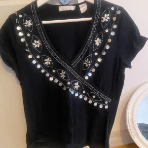 Svart omlottopp med paljettdetaljer - Svart omlottopp med v-ringning, korta ärmar och dekorativa paljetter samt broderade blommor längs halsringningen. Toppen har en unik och festlig design med glittrande detaljer som fångar blicken. Finns inga defekter. Passar storlek s-m
