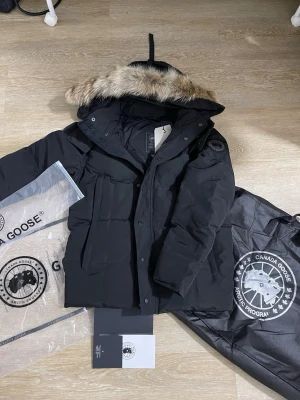 Svart parkas från Canada Goose - Säljer en svart parkas från Canada Goose med avtagbar huva och äkta pälsdetalj. Jackan har flera stora fickor, diskret logotyp på ärmen och är fodrad för extra värme. Perfekt för kalla vinterdagar och riktigt snygg streetstil.