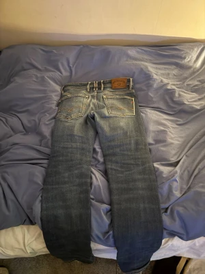 Replay jeans  - Säljer ett par klassiska blå jeans från replay  med raka ben och snygga slitningar. Jeansen har fem fickor, synliga knappar i gylfen och en brun läderpatch bak i midjan. Perfekta för en avslappnad och cool stil.