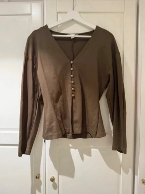 Grön blus med blommiga knappar - Stilren grön blus med v-ringning och långa ärmar. Framsidan har dekorativa små blommiga knappar i guld som ger en fin detalj