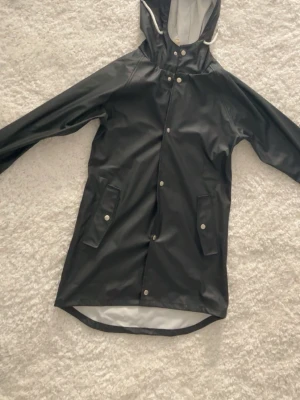 Svart regnjacka med huva - Stilren svart regnjacka med huva och tryckknappar framtill. Jackan har två stora fickor med lock och silverfärgade detaljer. Längre modell baktill och vitt innerfoder. Perfekt för regniga dagar och enkel att matcha med allt.