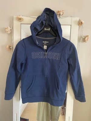 Blå hoodie från OshKosh - Mörkblå hoodie med dragkedja från OshKosh. Hoodien har huva, två fickor framtill och tryckt logga på bröstet. Tillverkad i mjukt bomullsmaterial, perfekt för en avslappnad stil.