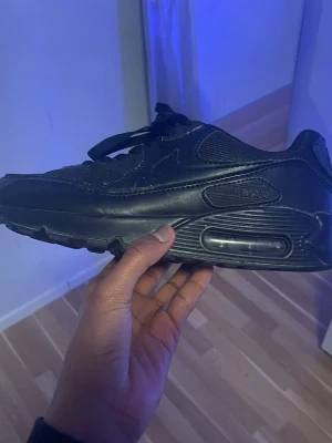 Nike Air Max 90 helsvart sneaker - Nike Air Max 90 i helsvart färg med klassisk chunky sula och synlig Air-enhet. Skon har snörning och är tillverkad i en mix av skinn och syntetmaterial för en clean och tidlös look. Perfekt för dig som gillar streetwear och vill ha en ikonisk sneaker.