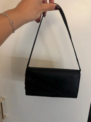 Satin Svart kuvertväska med axelrem - Snygg svart satin kuvertväska/clutch i avlång modell med dekorativa veck på framsidan. Är i använt skick men fortfarande väldigt snygg.