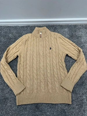 Beige kabelstickad tröja Polo Ralph Lauren - Kabelstickad beige tröja från Polo Ralph Lauren med halv dragkedja och hög krage. Tröjan har långa ärmar, ribbade muddar och nederkant samt den klassiska broderade loggan på bröstet. Perfekt för lager på lager och en stilren look.