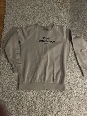 Beige sweatshirt från Peak Performance - Snygg beige sweatshirt från Peak Performance med svart broderad logga på bröstet. Tröjan har rund halsringning, ribbade muddar vid ärmslut och nederkant. Har inte använt den så många gånger. Stolek 150 