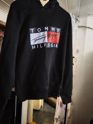 Svart hoodie från Tom - Svart hoodie från Tom  med stor logga framtill i vitt, rött och blått. Klassisk känguruficka och huva med dragsko. Mjuk bomullsblandning och avslappnad passform, perfekt för en chill och stilren look.