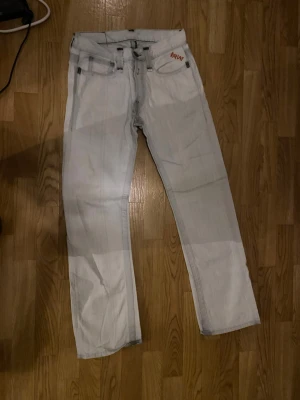 Ljusgrå raka jeans från Replay - Säljer ett par ljusgrå jeans från Replay med raka ben och klassisk femficksmodell. Jeansen har Replay-loggan broderad i rött på myntfickan och är tillverkade i denim. Perfekta för en avslappnad och trendig look.