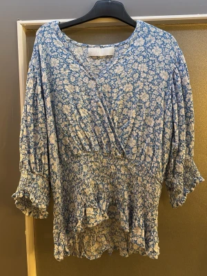 Blommig blå blus med trekvartsärm - Supersöt blommig blus i blått och vitt. Blusen har omlott-look framtill, smockad midja och volangkant nedtill. Trekvartsärmar med smock och volang vid ärmslut. Perfekt för en avslappnad men ändå chic stil.